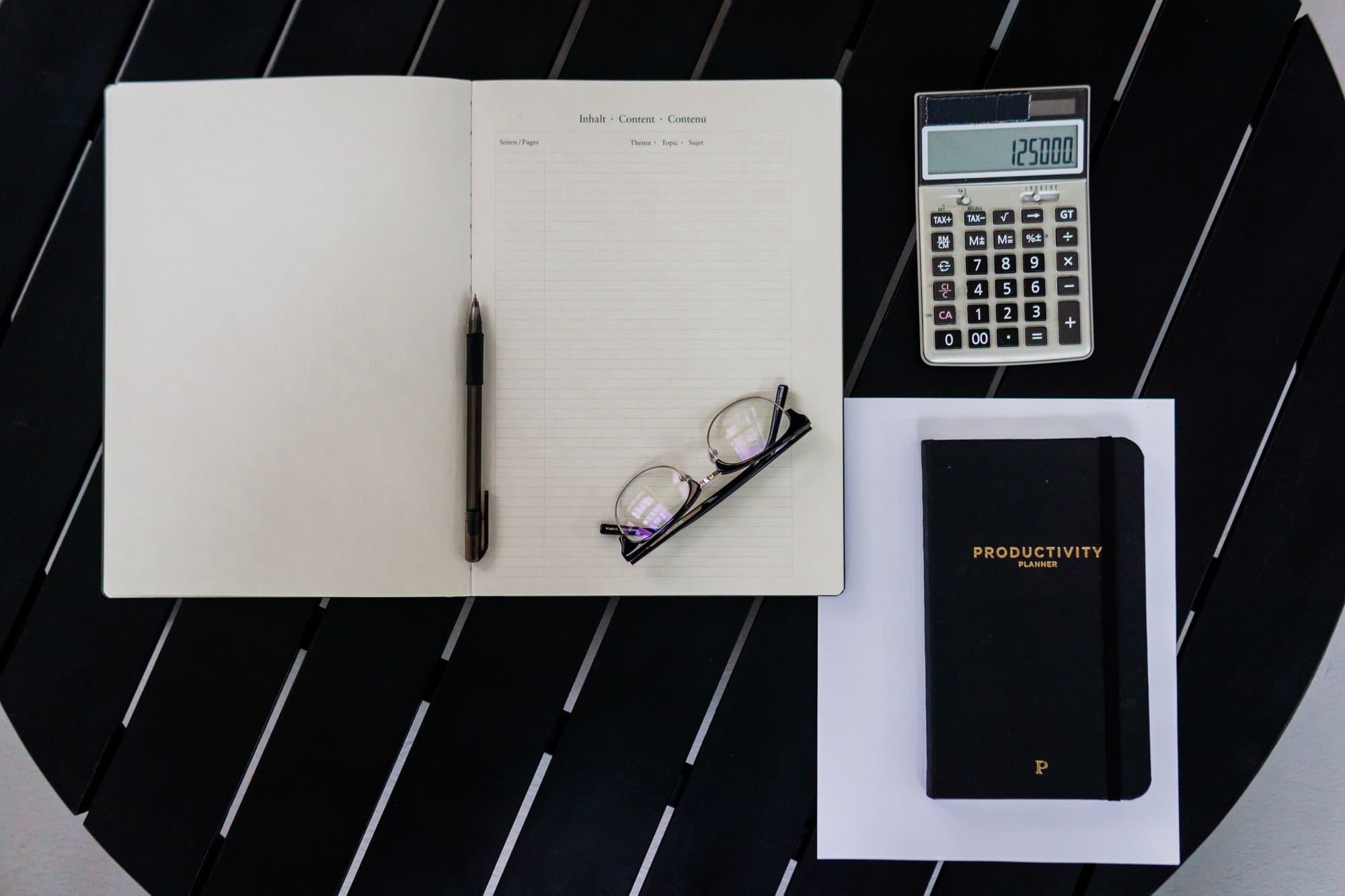 Open notebook, calculator, pen and glasses arranged on a dark desk
